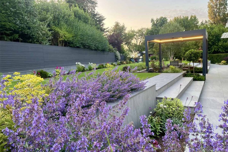 Patio featuring pergola has porcelain paving for garden tiles and steps leading up to lawn, surrounded by dark ash composite battens.