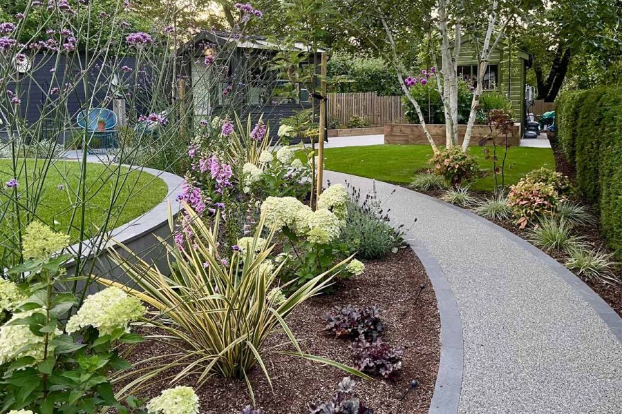 Curved pebble path with platinum grey porcelain setts lining either side runs through flowerbeds and bushes leading to garden shed.