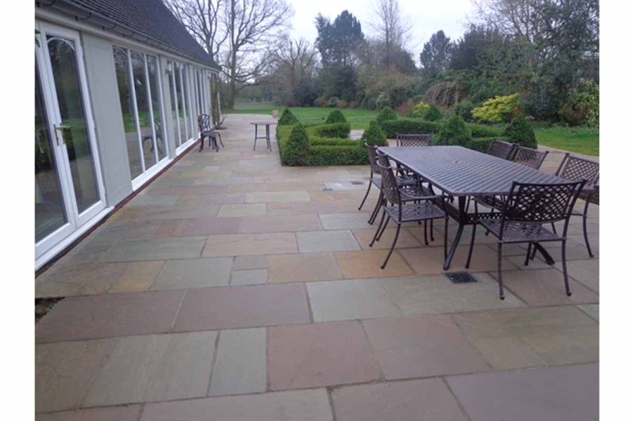 Very long patio of Tumbled Raj Green sandstone paving runs along back of house with multiple floor-to-ceiling windows.  