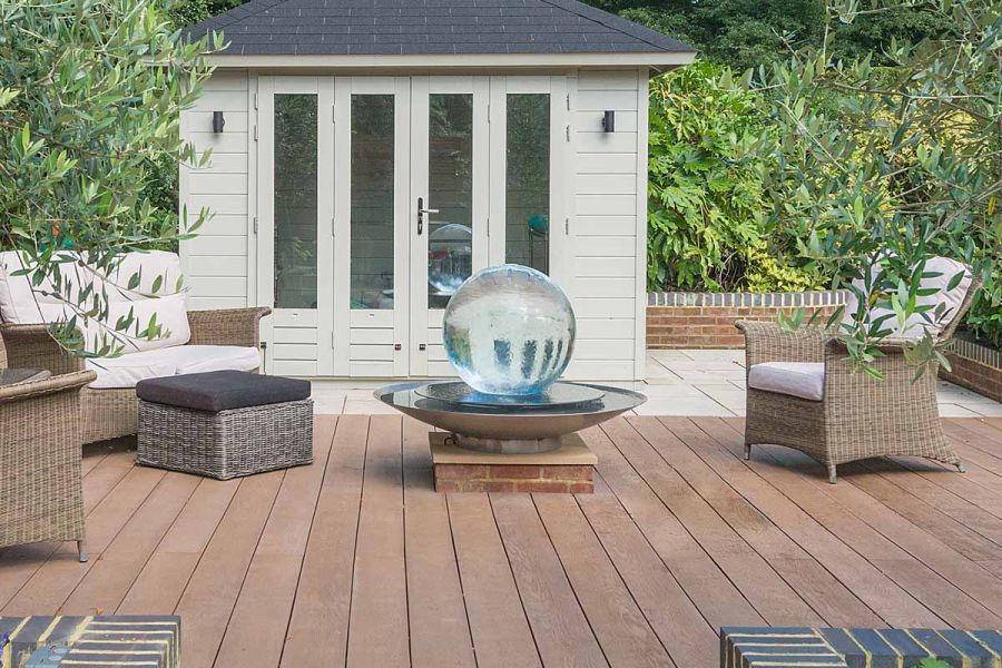 Spherical water fountain sat in the middle of outdoor furniture on coppered oak millboard decking surrounded by olive trees.