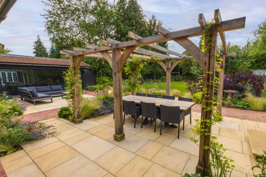 Climbing plants creep up wooden pergola next to clay paver path, Harvest Sawn sandstone patio host to dining table.