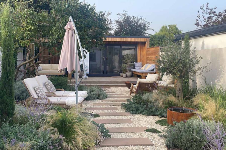 View of garden studio on decking area leads from buff sawn sandstone paving used as stepping stones surrounded by pebbles and planting.