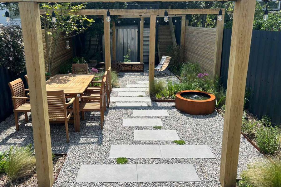 Slabs of Steel Grey Porcelain Paving with pebbles around create stepping stones through pergola, water feature and dining table either side.