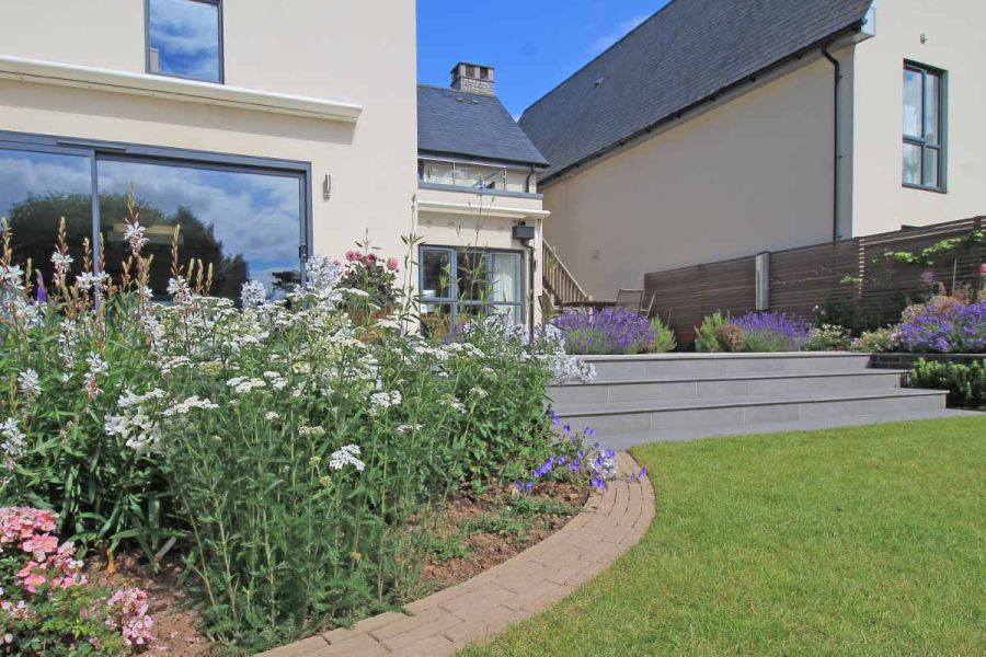 White house with french doors has patio leading down with steel grey porcelain 20mm bullnose steps towards lawn and curved flowerbeds.