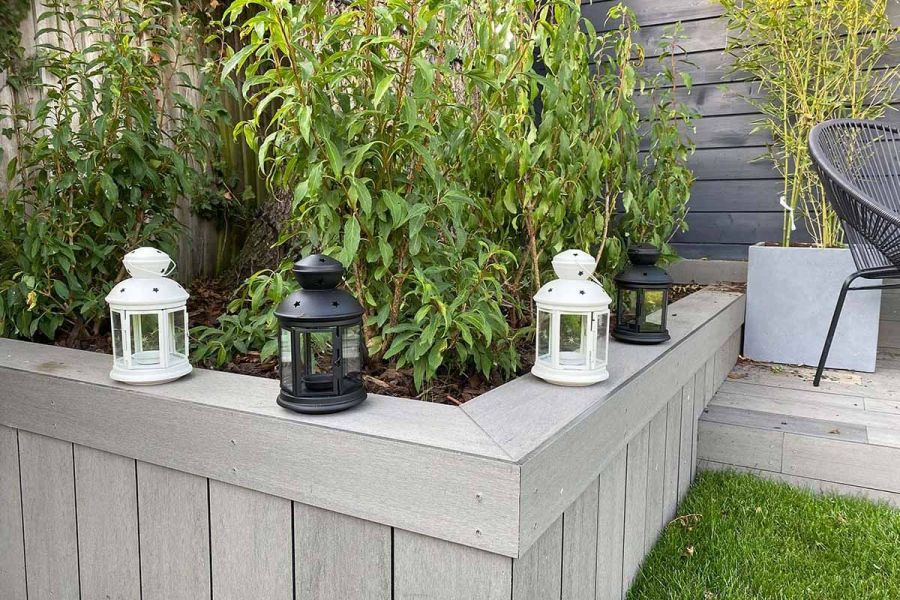 Raised bed with tall shrubs, bordered by Pebble Grey Brushed composite fascia boards, accented by white and grey lanterns along the edge.