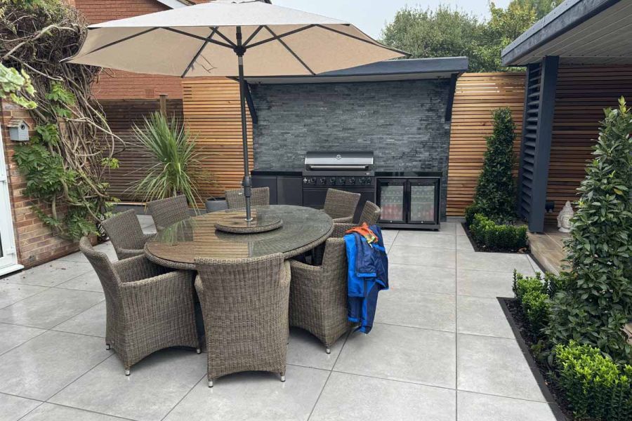 Outdoor dining set with umbrella sits on polvere large porcelain paving slabs, with slate feature wall used as BBQ splashback in the background.