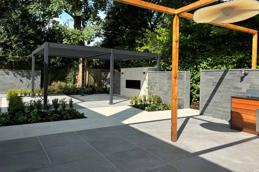 one wooden pergola and one metal pergola stand on anthracite and florence grey porcelain paving with sunlight casting shadows.