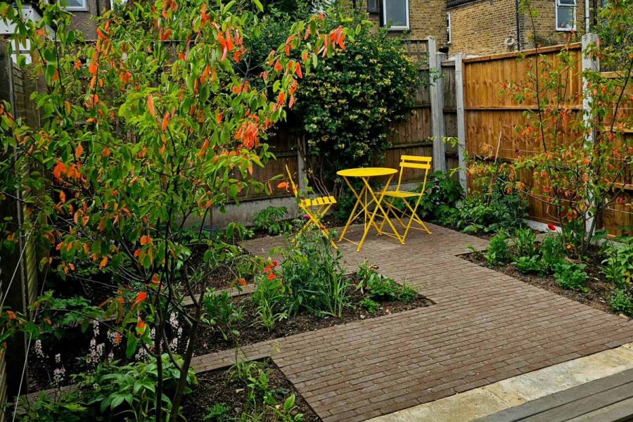 Aldridge clay paver patio in a J-shaped pattern has yellow metal furniture sitting on it with sunken flowerbeds surrounding.