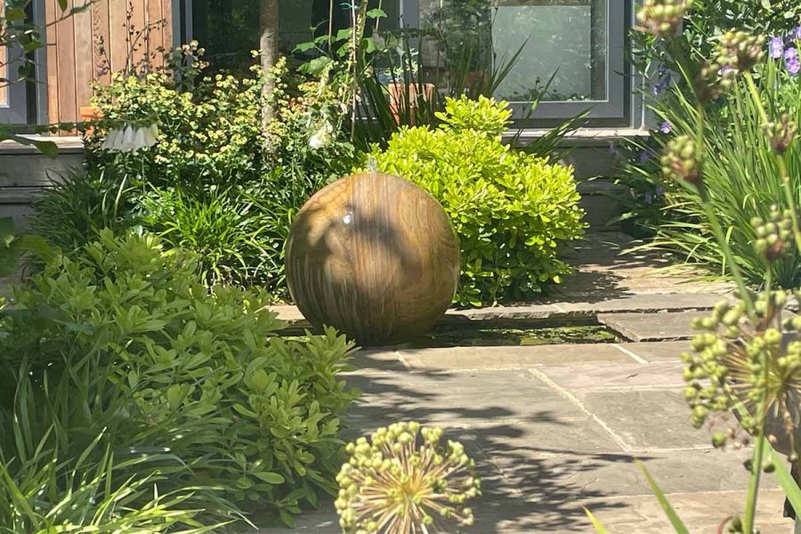 Sphere-shaped water feature made of stone sits on pond next to raj green indian sandstone patio surrounded by lush planting.