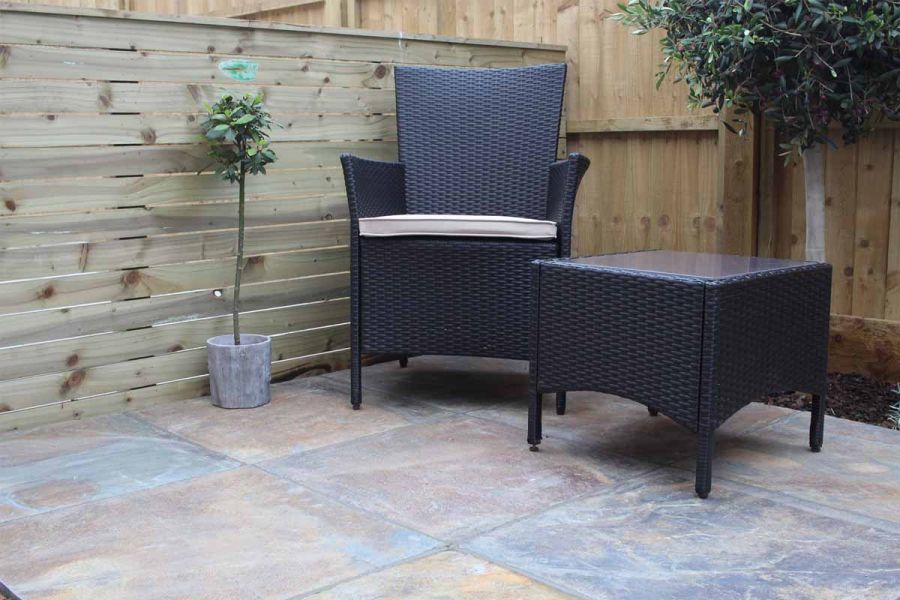 Single rattan armchair with cream cushion sits next to matching glass topped table, on top of rustic slate paving.