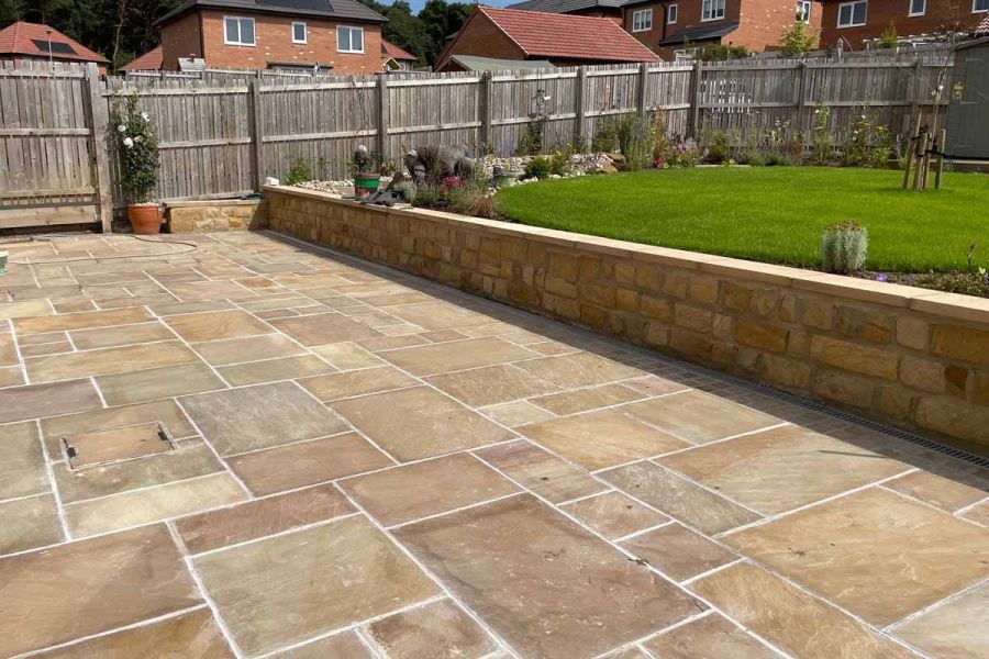 Patio of camel dust indian sandstone with white pointing is separated by stone wall with elevated lawn behind.