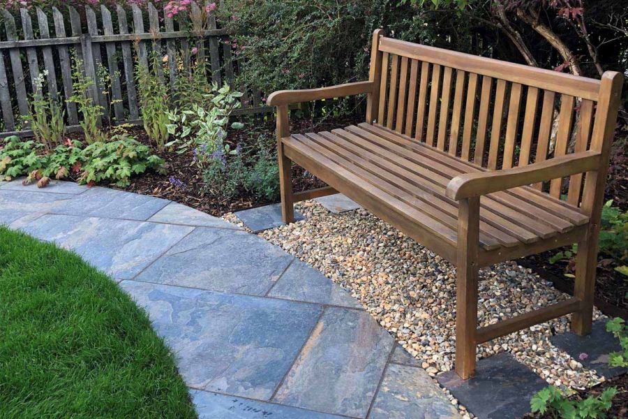 Wooden bench legs sit on 4 different slabs, surrounded by pebbles, path of rustic slate paving curves around lawn.