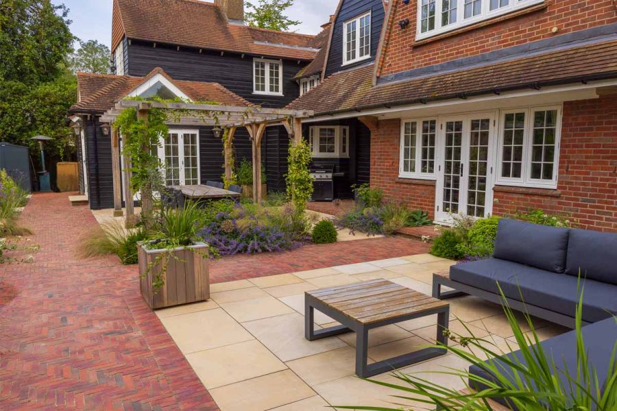 Clay Pavers with harvest sandstone patios inset, one host to lounge furniture and one to a dining table under wooden pergola.