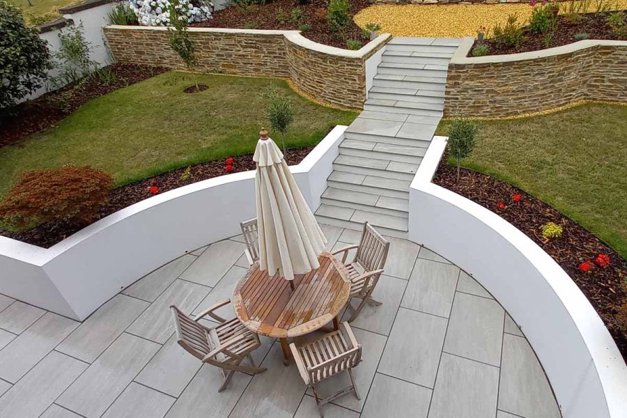 Circle patio of kandla grey porcelain paving with wooden dining table has steps leading to elevated lawn area bordered by flowerbeds.