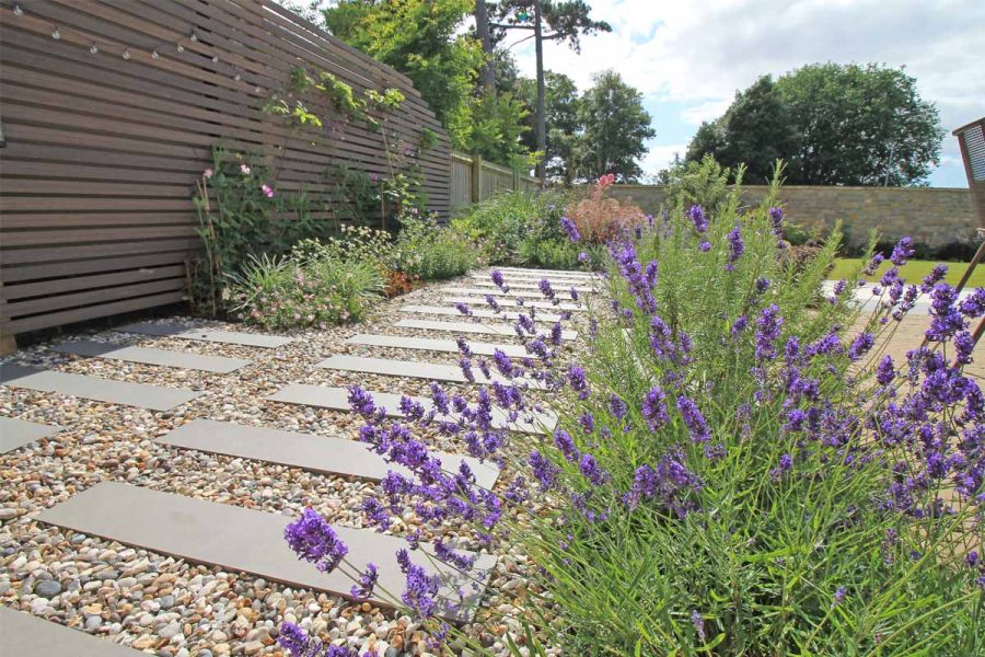 Close up view of steel grey porcelain planks inbetween light pebbles with flowerbeds and lawn surrounding.