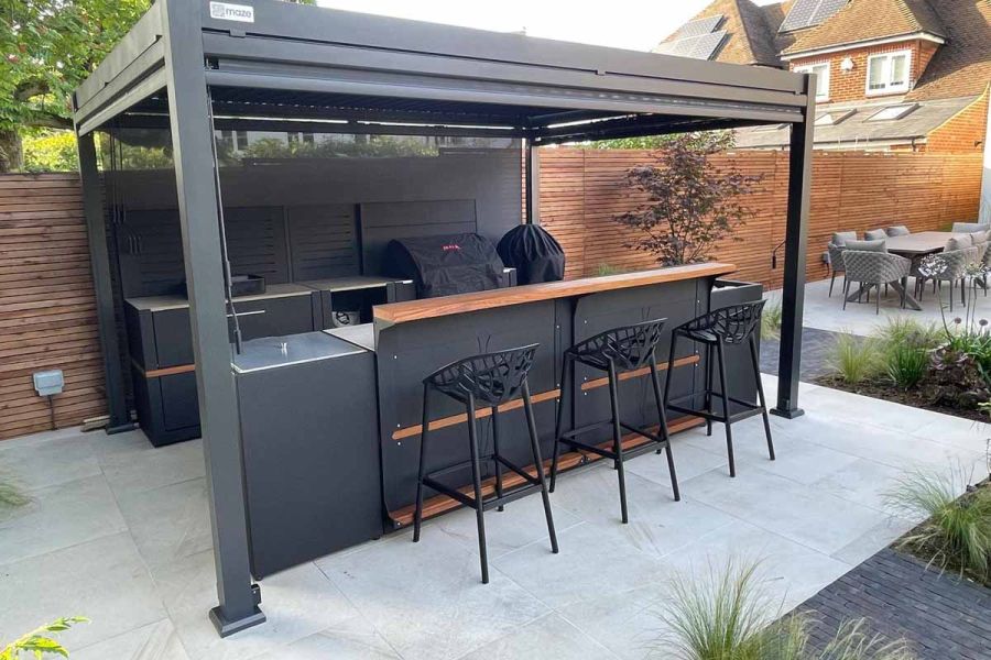 Three barstools sit under metal pergola on ash beige porcelain paving with insets of clay pavers and planting.