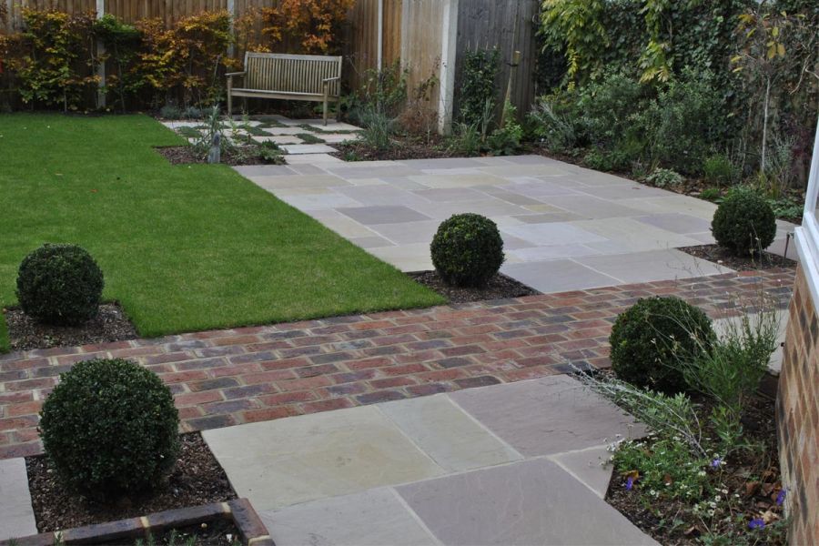 Clay paver path edged with topiary balls separates 2 paved areas of Tumbled Raj Green sandstone paving slabs. 