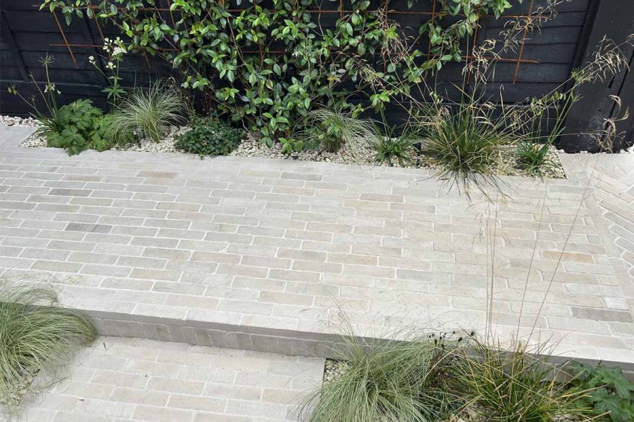 Path of beige sandstone patio bricks in 2 levels runs down length of bush with hedges planted on the steps below.