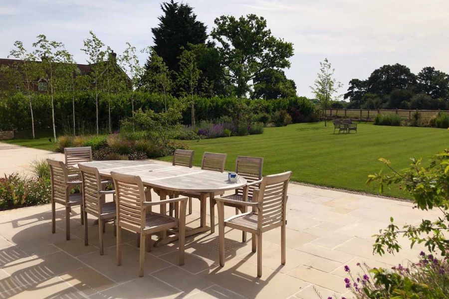 Wooden dining table sits on antique cream sandstone patio with views of large lawn bordered by hedges.