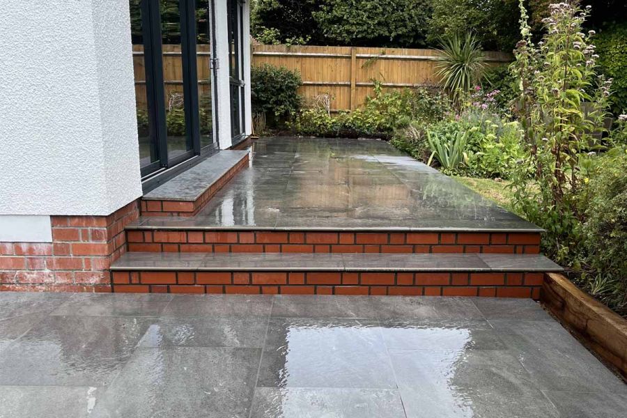 Multi-level patio features Platinum Grey paving and 20mm bullnose steps, complemented by white house walls and red brick-rendered steps.