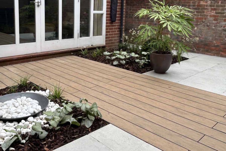 wide strip of warm teak brushed composite decking runs through grey porcelain paving with decorative pebbles and planting either side.