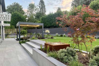 Porcelain patio featuring Proteus Grey 3x4 Aluminium Pergola with Optional Sides has porcelain paving for garden tiles and steps leading up to lawn.***The Landscape Design Studio
