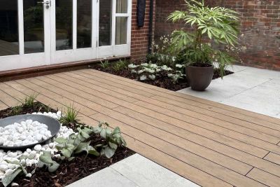 wide strip of warm teak brushed composite decking runs through grey porcelain paving with decorative pebbles and planting either side.***Designed by Chris Wild, www.cwgardendesign.co.uk | Built by TN1 Project Management, www.tn1projectmanagement.com