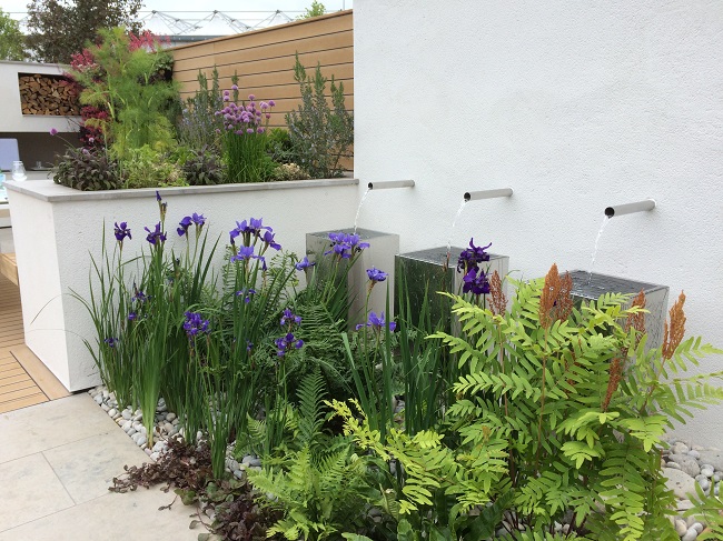 3 narrow pipes in white wall send water into 3 tall planters with irises and ferns in foreground. 