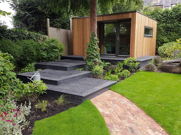 Path leads to Millboard Weathered Oak Embered platform steps rising to garden office under tall tree.