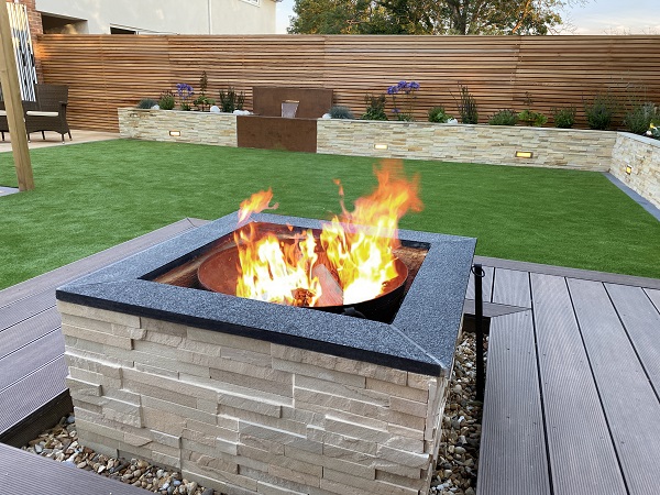 Square fire pit in middle of Chestnut Grooved composite decking in grassed garden with raised beds of Mint sandstone cladding.