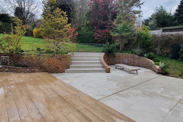 Patio of decking and Area 800x800 porcelain outside tiles, with curved flight of steps up to lawn with mature shrubs.