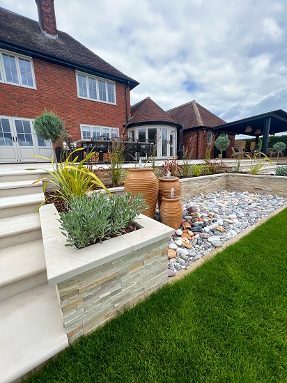 Raised bed faced with Mint sandstone cladding makes retaining wall to large raised patio.