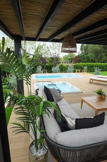 Sofa under pergola with view over hot tub and swimming pool to boundary hedge.