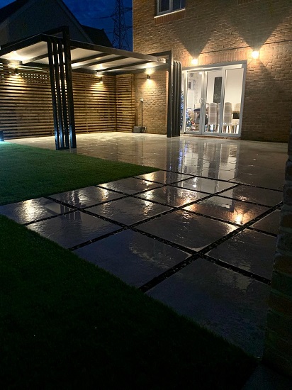 Square paving slabs with wide joints reflect lights from back of house in dark garden. 