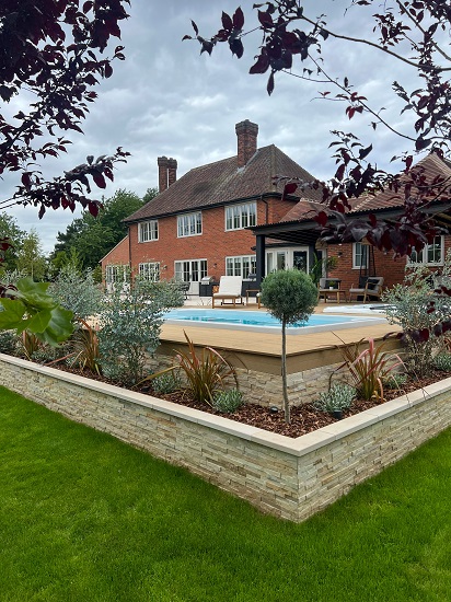 Corner of large raised patio with surrounding retaining bed faced with Mint sandstone cladding. Swimming pool beyond.