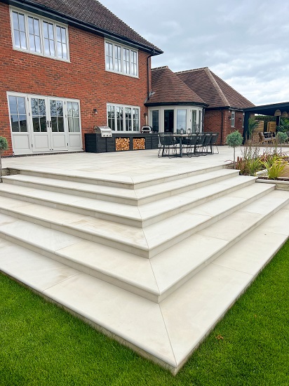 Corner of Beige sawn sandstone raised patio, wrapped around brick house, with bullnose steps descending to lawn.