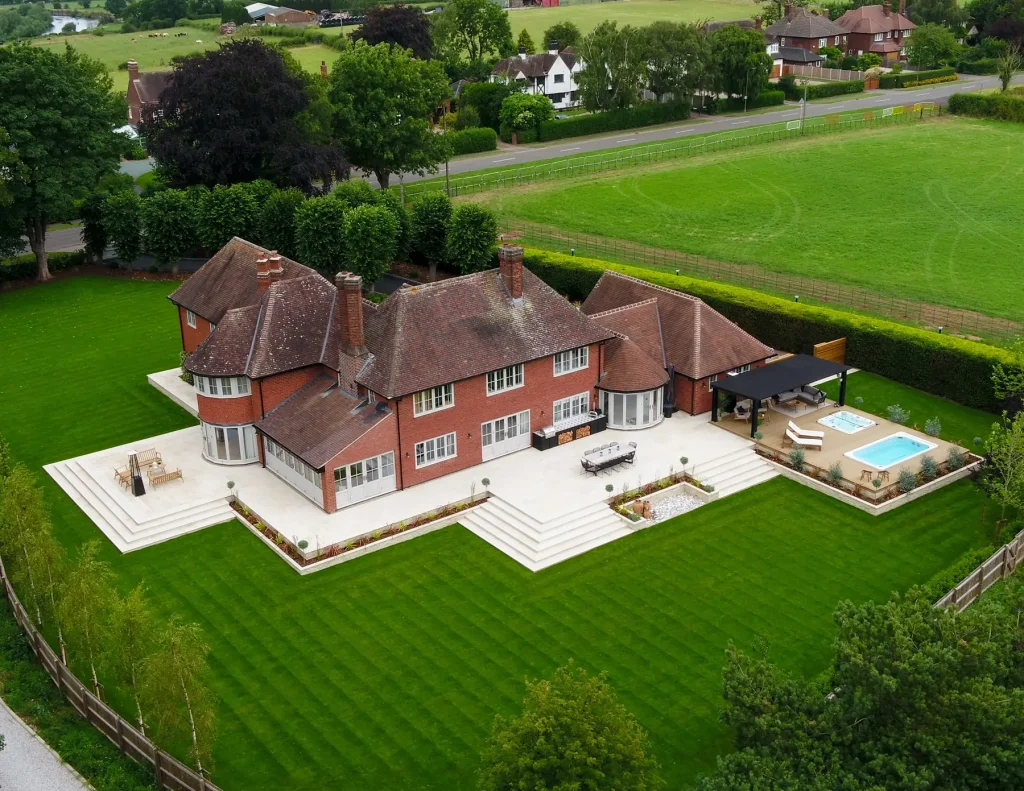 Drone view of large house with Beige sawn sandstone wrap-around patio, swimming pool and huge lawn.