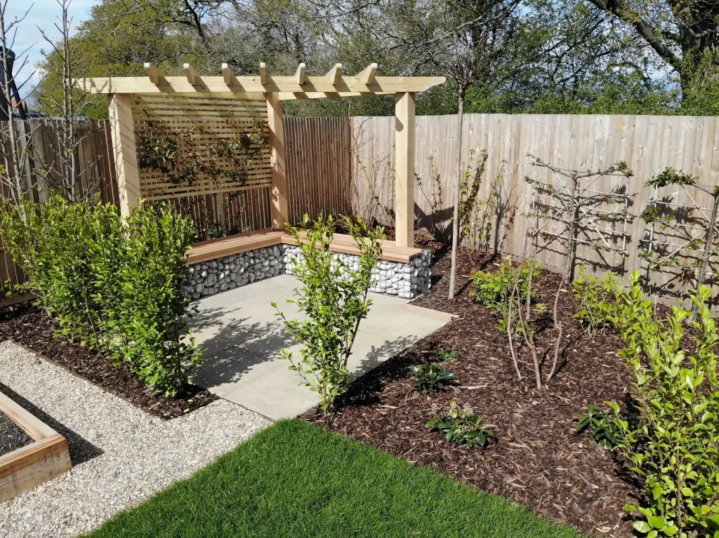 Triangular wooden pergola anchored in wood and gabion L-shaped bench on square stone patio in fenced corner of garden.