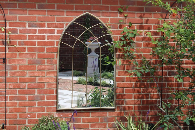 A gothic arch mirror on brick wall reflects garden with Venetian Beige paving and urn.