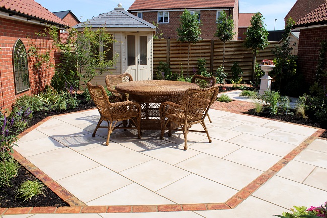 Garden dining set sits on Venetian Beige porcelain paving outlined by single course of Cotswold clay pavers. Summerhouse behind. 