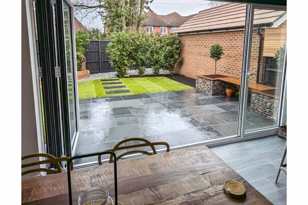View through open bifold doors across deep Brazilian Black slate patio to lawn with straight path towards hedge.