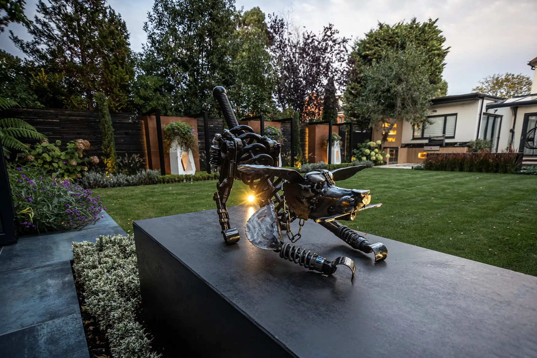 Metal caricature of dog on plinth faced with Steel Dark DesignClad in garden with large lawn and 3 large white planters against fence in background.