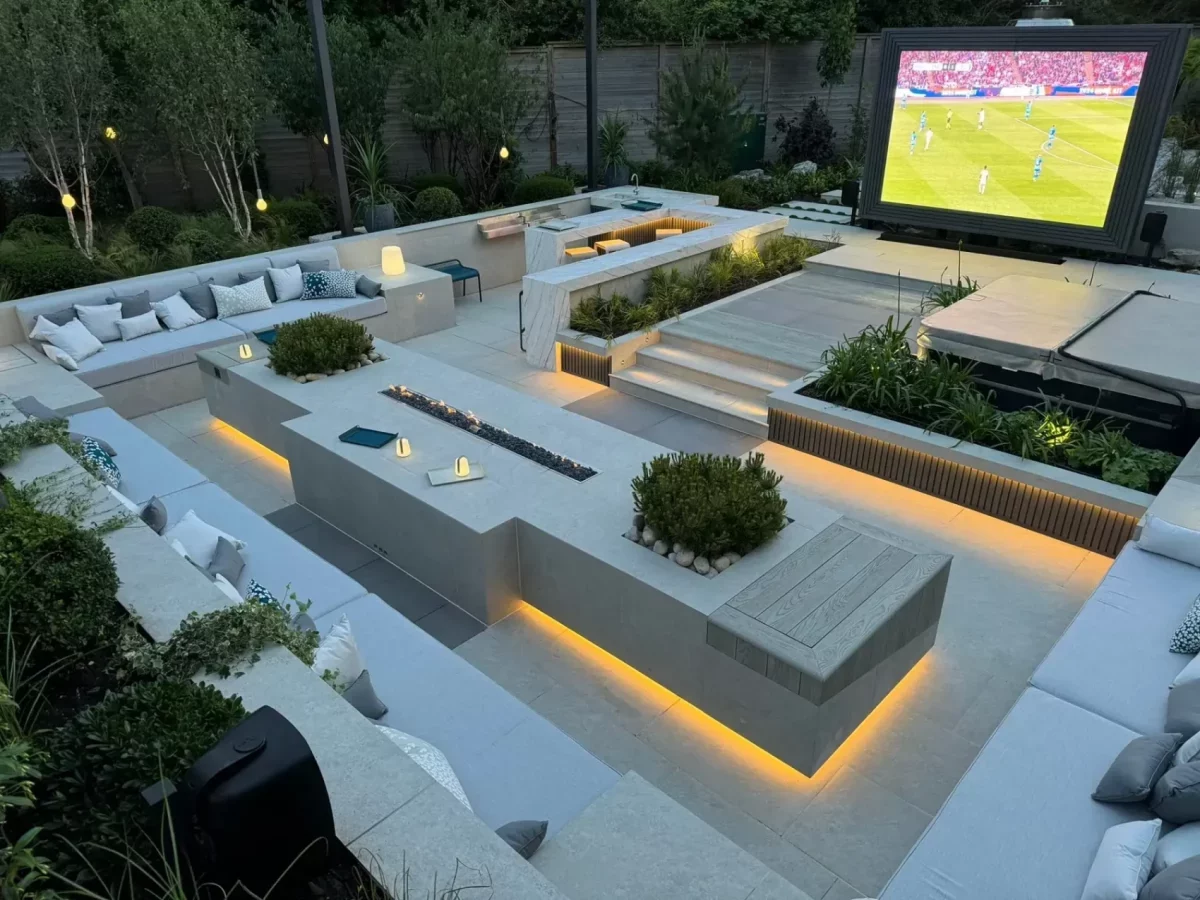 Large sunken seating area with sofas on 3 sides and long firepit table, in party garden with large TV screen