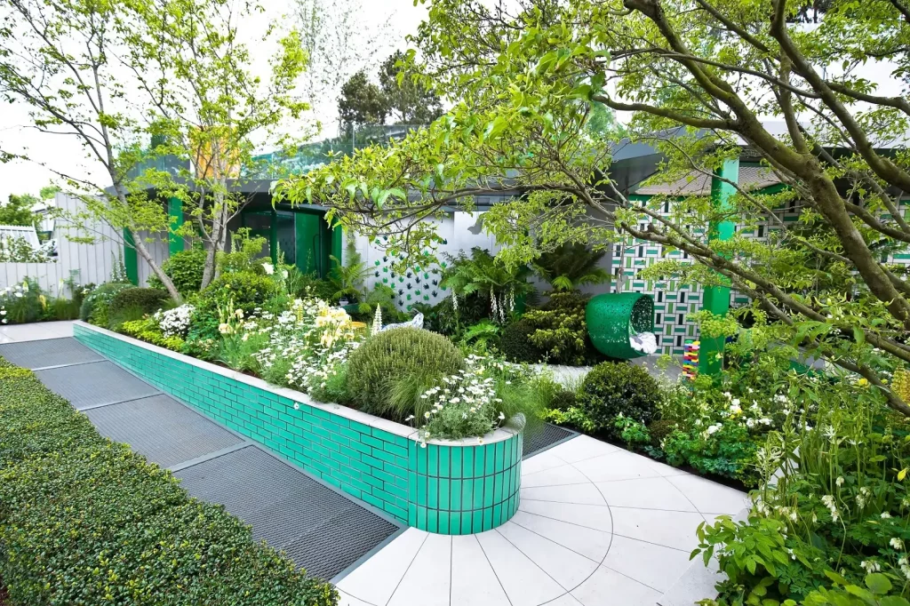 Accessible garden with smooth path making hairpin turn around green tiled raised bed. Pavillion in background. 